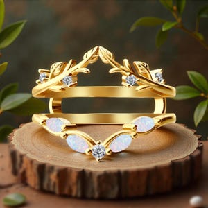 May include: A gold-colored ring guard with a leaf design and white opal accents. The ring guard features small, round, clear gemstones and is displayed on a wooden surface.