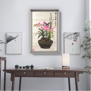 May include: A framed watercolor painting of a potted orchid with pink flowers and green leaves. The painting is in a gray frame and is displayed on a white wall. Two other framed paintings are also on the wall.