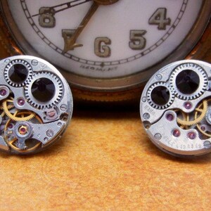 May include: A pair of silver-toned cufflinks with intricate clockwork designs. Each cufflink features a black face with gears and a small black jewel.