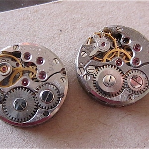 May include: Two silver watch movements with gold gears and red jewels. The movements are circular and have a diameter of approximately 1 inch.