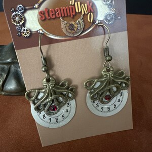 May include: Steampunk style earrings with a clock face and an octopus design. The clock face has a red rhinestone in the center and the numbers 1 through 12 around the edge. The octopus design is made of metal and has a detailed design.