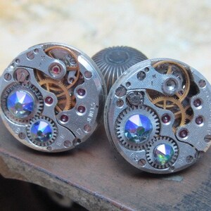 May include: A pair of silver cufflinks with a steampunk design. Each cufflink features a circular watch face with exposed gears and two iridescent blue rhinestones.