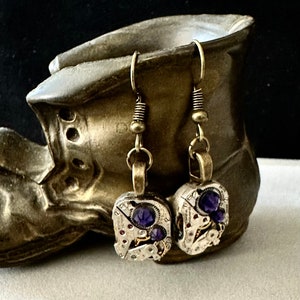 May include: A pair of steampunk earrings with purple gemstones set in antique watch parts. The earrings are hanging from bronze hooks.