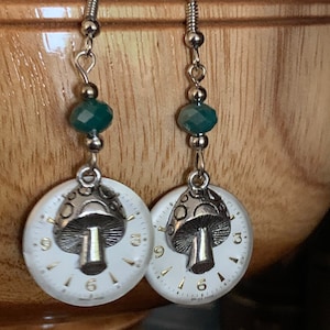 May include: Silver-toned earrings featuring white watch faces with gold numerals and a silver mushroom charm. Each earring has a faceted teal bead and a silver hook. The earrings are a unique, handcrafted accessory.