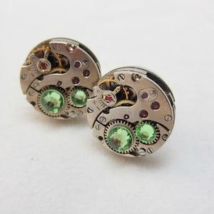 May include: A pair of circular cufflinks crafted from vintage watch movements. Each piece features exposed silver-toned gears, small metallic screws, and two round, faceted light green gemstones set within the intricate mechanical clockwork components.