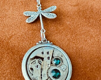 May include: Silver necklace with a dragonfly pendant and a round watch face pendant with green gemstones. The watch face pendant has visible gears and other watch parts.