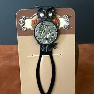 May include: Black steampunk owl bookmark with a clockwork center and iridescent gem eyes. The bookmark features the word "Steampunk" on the card. The owl is attached to a black, teardrop-shaped bookmark.