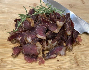 May include: A close-up of a pile of dried, seasoned meat strips on a wooden cutting board. The meat is dark red and brown, and there are sprigs of rosemary scattered throughout.