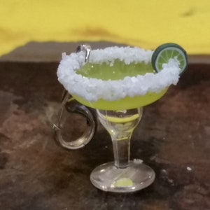 May include: A miniature margarita glass keychain with a lime wedge garnish and a salted rim. The glass is filled with a green liquid.