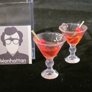 May include: A pair of red cocktail earrings with a cherry and a toothpick. The earrings are shaped like martini glasses. The earrings are in front of a card with the word "Manhattan" on it.