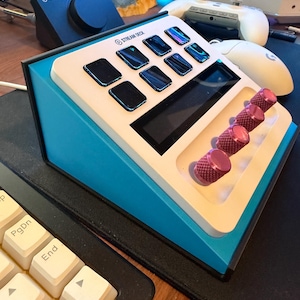 May include: A Stream Deck controller featuring a white faceplate with eight black square buttons, a horizontal display screen, and four pink textured rotary knobs. The device has a bright blue angled side casing and a black base frame.
