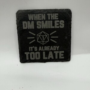 Puede incluir: Un posavasos cuadrado de pizarra negra con el texto "WHEN THE DM SMILES, IT'S ALREADY TOO LATE" en gris. Un gráfico estilizado de dado D20 está centrado debajo de la primera línea. Para los entusiastas de los juegos de mesa.