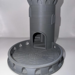 May include: A gray, 3D-printed castle tower with a circular base and a spiral staircase visible through an arched doorway. The tower has a crenelated top and arched windows. The base has a decorative, arched railing.