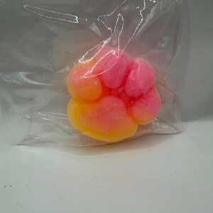 May include: A paw-shaped bath bomb in a clear plastic bag. The bath bomb has a gradient of colors, transitioning from yellow to orange to pink. The product is likely for use in a bath.