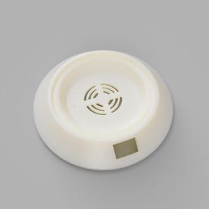 May include: A cream-colored, circular object with a recessed center and a series of small, curved slots. The object has a flat base and a slightly raised rim. A small, rectangular indentation is visible on the side.