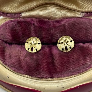 May include: A pair of gold stud earrings shaped like sand dollars, featuring a textured surface and a central star-like pattern. The earrings are displayed inside a vintage-style, heart-shaped jewelry box lined with deep purple velvet fabric.