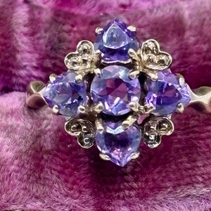 May include: A silver ring with a cluster of purple gemstones. The ring features heart-shaped details and a central arrangement of round and pear-shaped stones. The gemstones are a vibrant purple, set against a silver band, showcasing a detailed design.