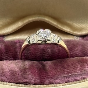 May include: A vintage engagement ring with a central square-cut diamond and smaller accent diamonds. The ring has a gold band and is set in a white metal setting. It rests in a purple velvet ring box.