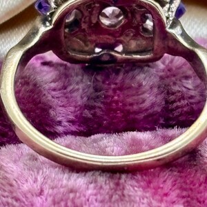 May include: Close-up of a vintage ring with a gold band. The ring features a central clear stone surrounded by smaller clear stones and vibrant purple stones. The ring is set against a plush, purple background.