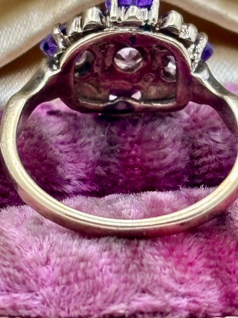 May include: Close-up of a vintage ring with a gold band. The ring features a central clear stone surrounded by smaller clear stones and vibrant purple stones. The ring is set against a plush, purple background.