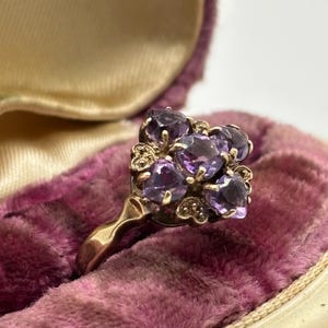 May include: A gold ring with a cluster of oval-cut purple gemstones. The ring is set against a backdrop of a vintage, plush, purple velvet jewellery box, with a cream-coloured interior.
