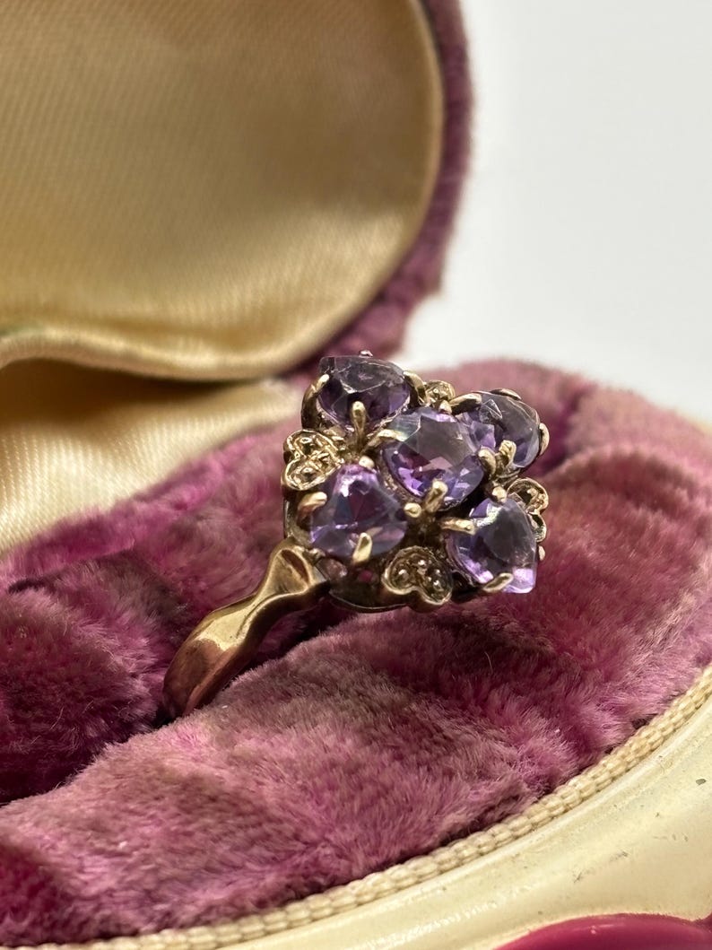 May include: A gold ring with a cluster of oval-cut purple gemstones. The ring is set against a backdrop of a vintage, plush, purple velvet jewellery box, with a cream-coloured interior.