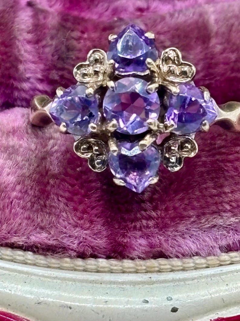 May include: A silver ring with a cluster of purple gemstones. The ring features heart-shaped details and a central arrangement of round and pear-shaped stones. The gemstones are a vibrant purple, set against a silver band, showcasing a detailed design.