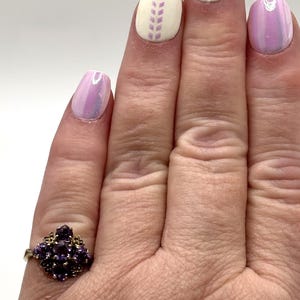 May include: A gold ring set with a cluster of small, dark purple gemstones. The hand shows manicured nails with a light purple iridescent polish and white nails with a lavender design.