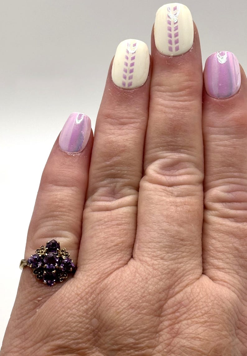 May include: A gold ring set with a cluster of small, dark purple gemstones. The hand shows manicured nails with a light purple iridescent polish and white nails with a lavender design.