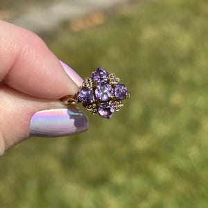 May include: A gold ring with a floral arrangement of purple gemstones. The ring has a central cluster of purple stones, surrounded by smaller accent stones. The ring is held up against a blurred green background, showcasing its intricate design and colour.