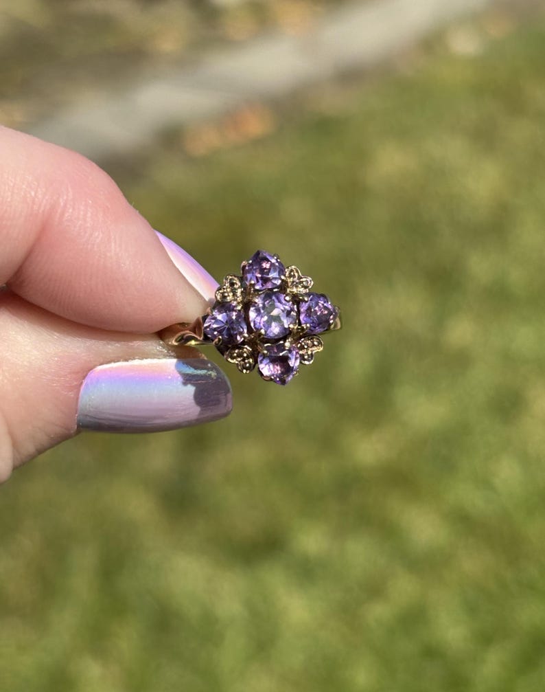 May include: A gold ring with a floral arrangement of purple gemstones. The ring has a central cluster of purple stones, surrounded by smaller accent stones. The ring is held up against a blurred green background, showcasing its intricate design and colour.