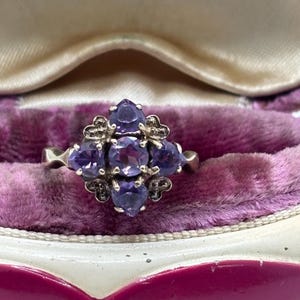 May include: A silver ring with a floral arrangement of purple gemstones. The ring has small decorative details. It is presented on a purple velvet surface within a jewellery box.