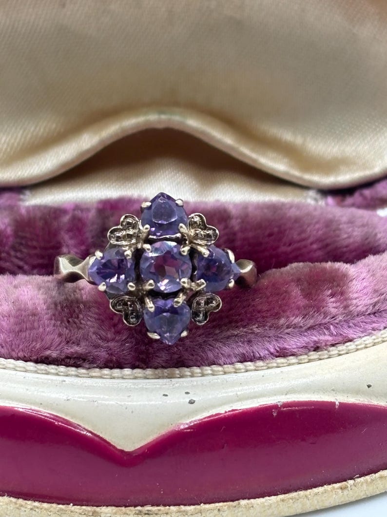 May include: A silver ring with a floral arrangement of purple gemstones. The ring has small decorative details. It is presented on a purple velvet surface within a jewellery box.
