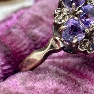 May include: Close-up of a vintage ring featuring multiple oval-cut, deep purple gemstones. The ring has an ornate, gold-toned setting with decorative filigree details. The ring's band is smooth and polished.