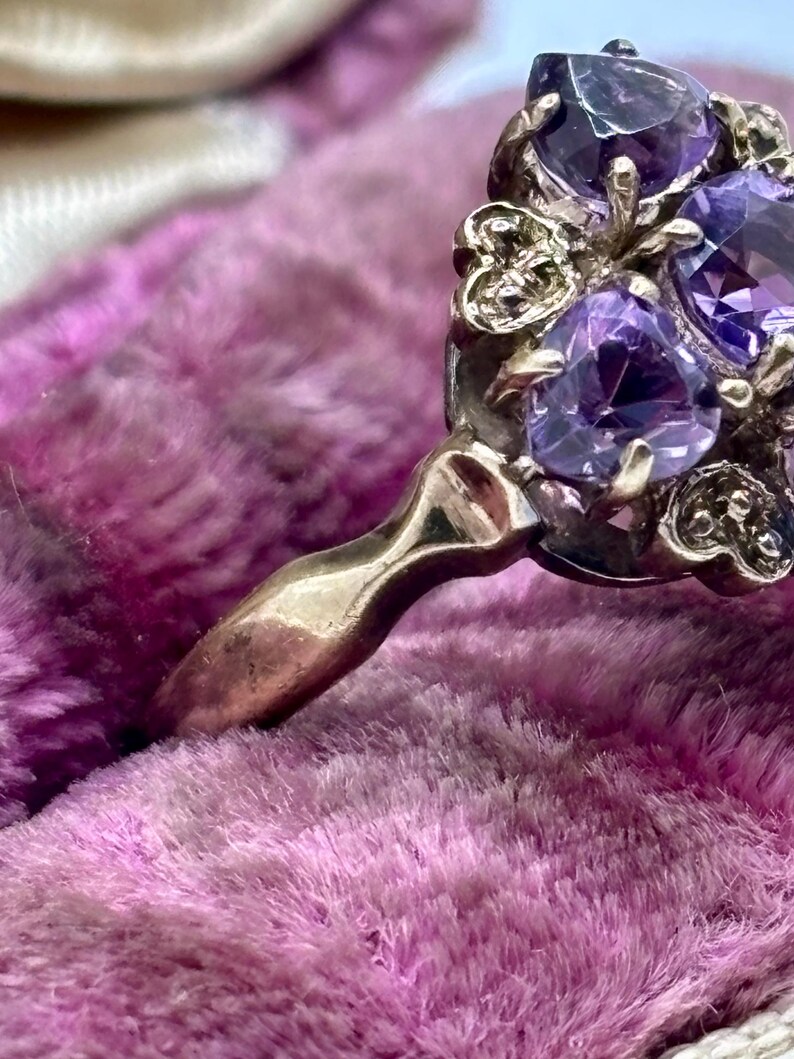 May include: Close-up of a vintage ring featuring multiple oval-cut, deep purple gemstones. The ring has an ornate, gold-toned setting with decorative filigree details. The ring's band is smooth and polished.