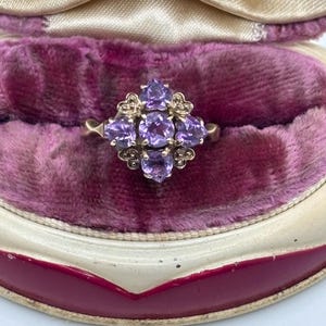 May include: A gold ring with a cluster of heart-shaped, purple gemstones. The ring is set in a floral design and presented in a vintage purple velvet jewellery box. The gemstones are arranged in a symmetrical pattern, creating a classic look.