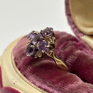 May include: A gold ring with a cluster of oval purple gemstones. The ring is set against a pink velvet background inside a vintage jewellery box. The gemstones are arranged in a floral pattern.