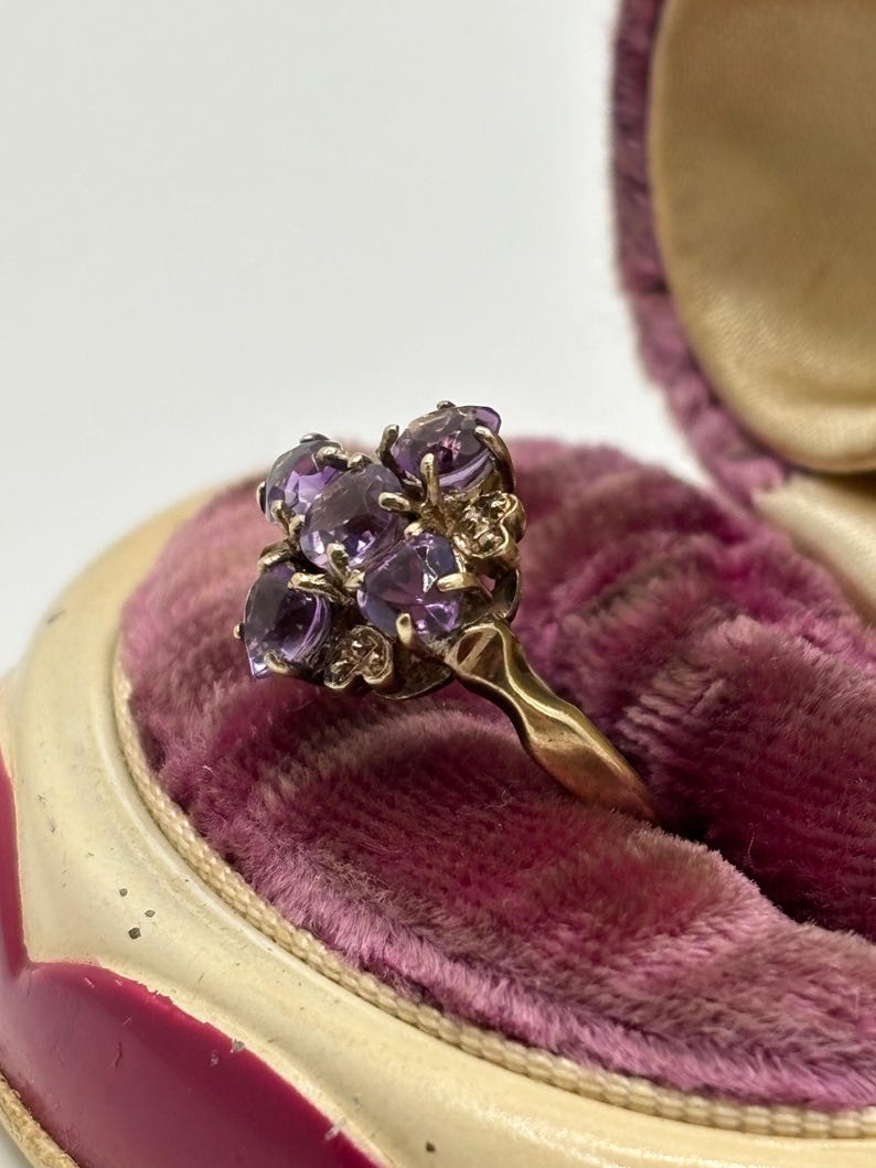 May include: A gold ring with a cluster of oval purple gemstones. The ring is set against a pink velvet background inside a vintage jewellery box. The gemstones are arranged in a floral pattern.