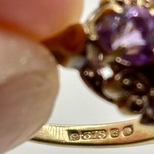 May include: Close-up of a gold ring featuring a large, oval, purple gemstone. The ring has an ornate, filigree design around the gemstone. The band is stamped with "9.375" and other markings.