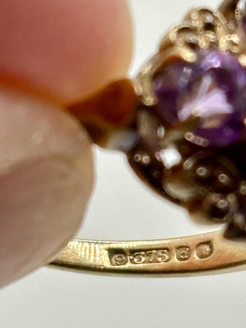 May include: Close-up of a gold ring featuring a large, oval, purple gemstone. The ring has an ornate, filigree design around the gemstone. The band is stamped with "9.375" and other markings.