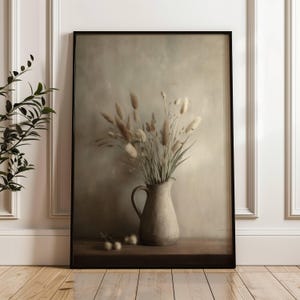 May include: A framed art print depicting a still life arrangement of dried flowers in a rustic ceramic jug. The artwork uses a muted colour palette of beige, brown, and green. The frame is black, and the print is displayed on a wooden surface.