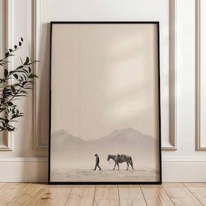 Pode incluir: Impressão emoldurada em tons sépia retratando uma pessoa caminhando ao lado de um cavalo selado em uma paisagem desértica. Montanhas são visíveis ao fundo sob um céu enevoado. A obra de arte está em uma moldura preta, adequada para decoração.