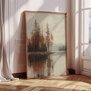 May include: A framed landscape painting depicts a serene lake scene with a cluster of trees in autumn colors. The artwork features a gold frame and is leaning against a white wall with a wooden floor.