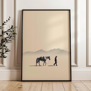 Pode incluir: Impressão de arte emoldurada com uma cena minimalista do deserto. Uma pessoa de chapéu caminha ao lado de um cavalo selado, com montanhas ao fundo. A obra de arte é em tons neutros, emoldurada em preto e sobre um fundo bege.