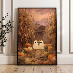 May include: A framed Halloween print depicting two white ghost figures in a cornfield with pumpkins. The artwork uses a warm colour palette of orange, brown, and yellow, with a rustic barn in the background. The print is in a black frame.