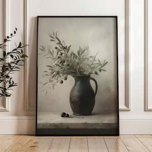 Könnte beinhalten: Gerahmter Kunstdruck mit einem Stillleben. Ein dunkler, rustikaler Krug enthält Olivenzweige mit grünen Oliven. Zwei Oliven liegen auf einer Steinoberfläche. Das Kunstwerk ist in Schwarz gerahmt.
