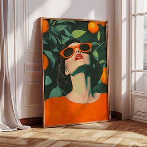 May include: A framed art print depicting a woman in orange sunglasses and top, gazing upwards. The background shows green leaves and oranges. The artwork has a gold frame and is propped against a white wall.