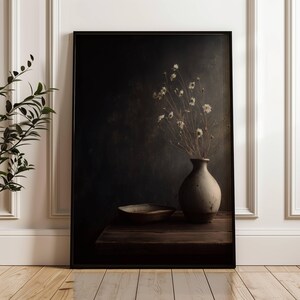 Pode incluir: Uma impressão emoldurada de uma natureza morta. Apresenta um vaso texturizado com flores secas e uma tigela pequena sobre uma superfície de madeira escura. O fundo é de uma cor escura e melancólica. A obra de arte é exibida contra uma parede branca.