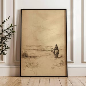 Pode incluir: Esboço em tons sépia emoldurado de um cowboy a cavalo atravessando uma paisagem desértica. A obra de arte apresenta um cowboy com chapéu e casaco, montando um cavalo por um campo com montanhas ao fundo. A moldura é preta.