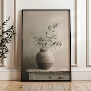 Puede incluir: Impresión enmarcada que presenta una naturaleza muerta. Un jarrón rústico de dos tonos contiene ramas de olivo, sobre un fondo neutro. La obra de arte se muestra sobre una superficie de madera clara y desgastada, enmarcada en negro.